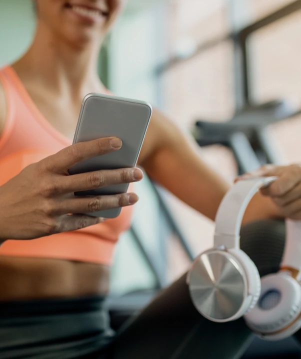 Femme souriante en tenue de sport tenant un téléphone intelligent dans une main et un casque d’écoute blanc dans l’autre, assise dans une salle d’entraînement lumineuse.