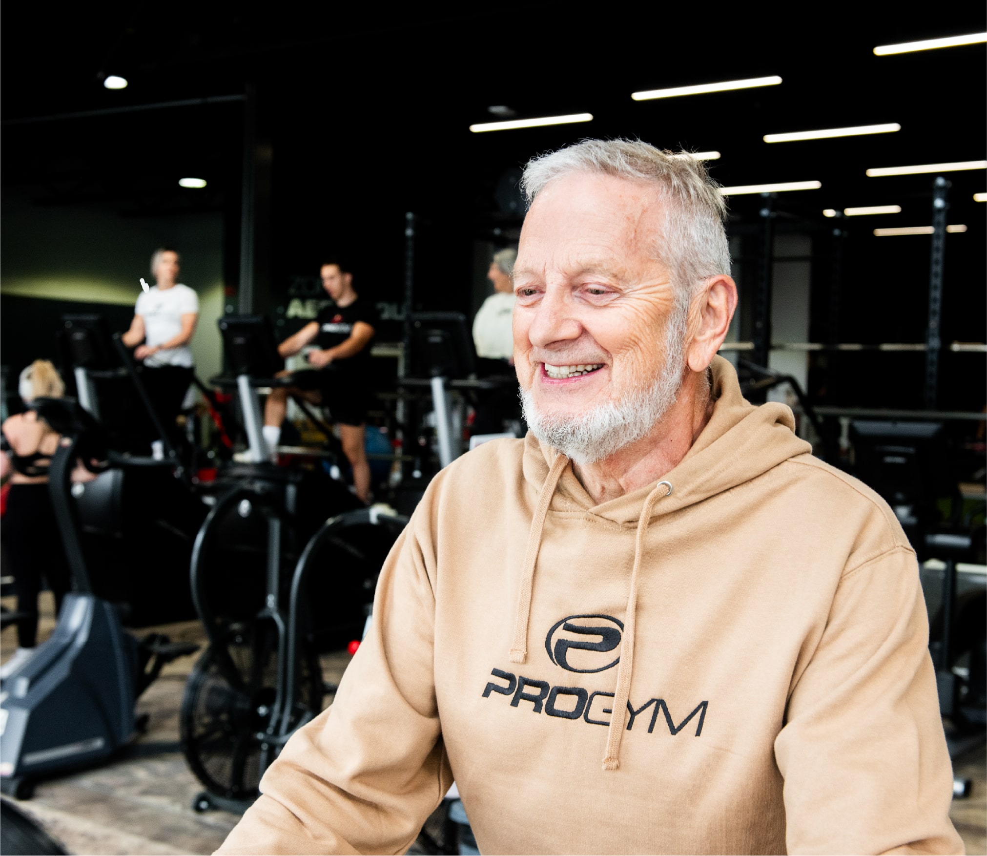 Homme souriant aux cheveux gris portant un hoodie beige ProGym, en plein entraînement dans une salle de sport moderne, avec d’autres membres visibles à l’arrière-plan.