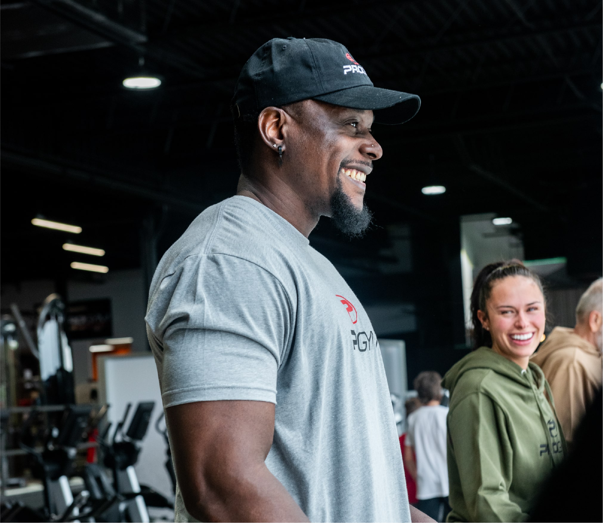 Homme souriant portant un chandail gris et une casquette noire ProGym, debout dans une salle d’entraînement, accompagné d’une femme en hoodie vert olive également souriante en arrière-plan.
