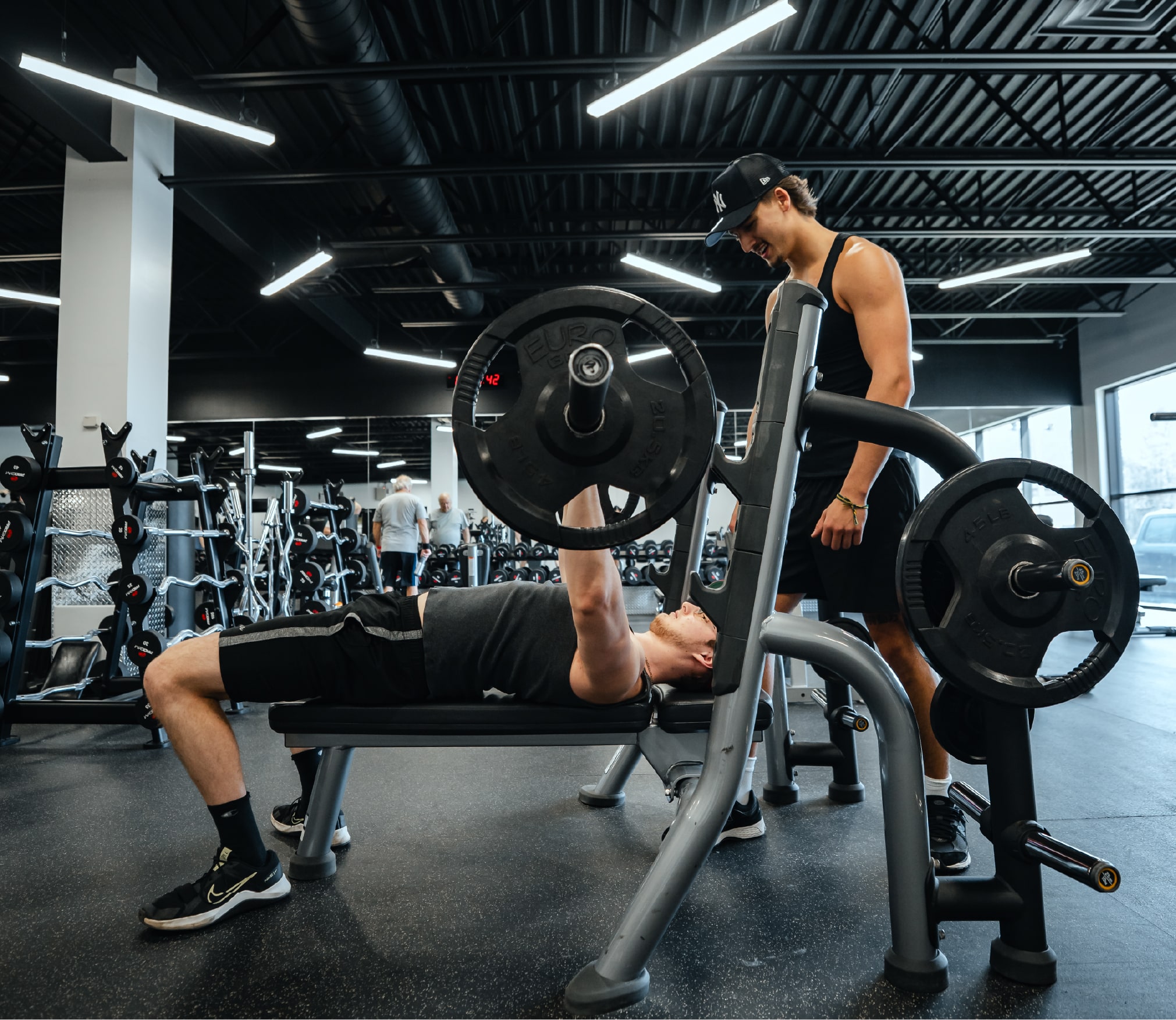 Entraînement de musculation dans un gym ProGym : un homme effectue un développé couché sous la supervision d’un partenaire, dans une salle moderne équipée d’appareils et de poids libres.