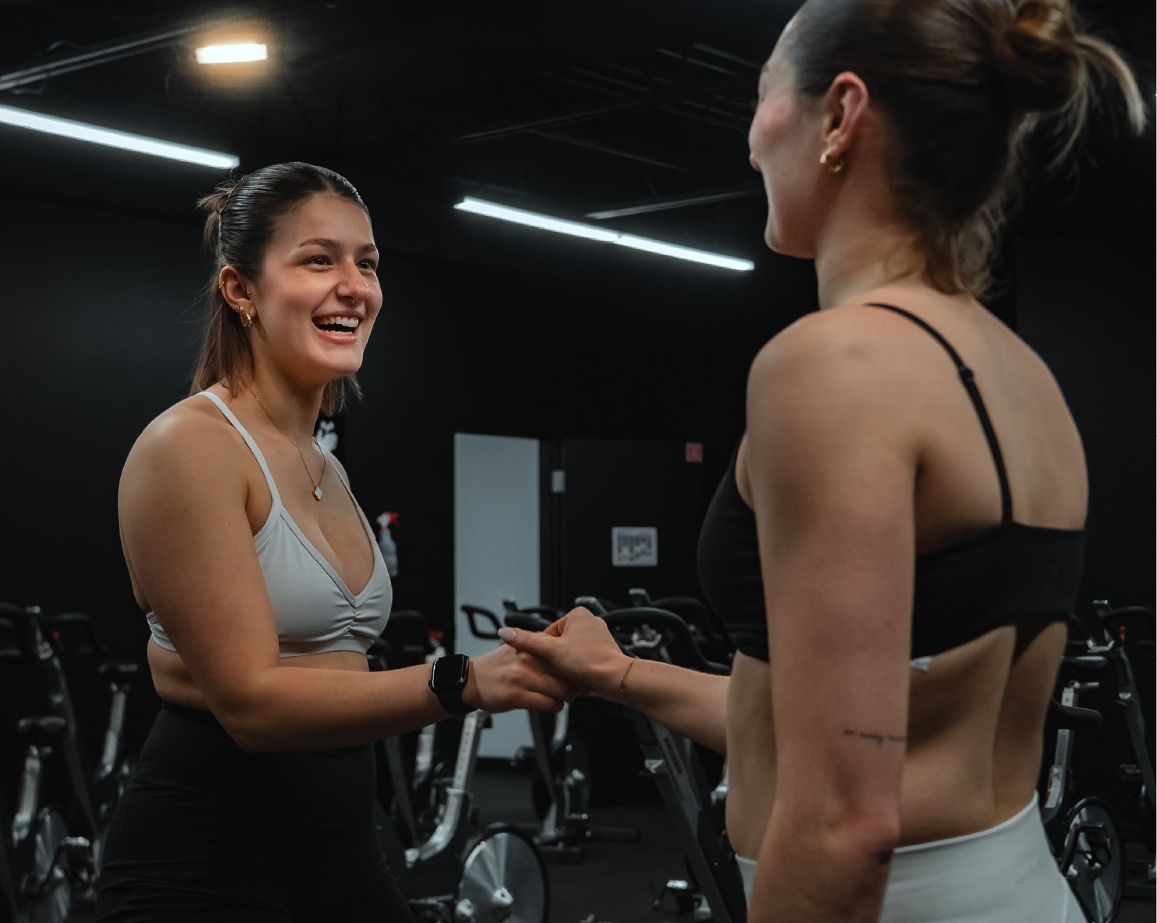 Deux femmes souriantes se serrent la main dans une salle de spinning, habillées en vêtements de sport, dans une ambiance conviviale au ProGym.