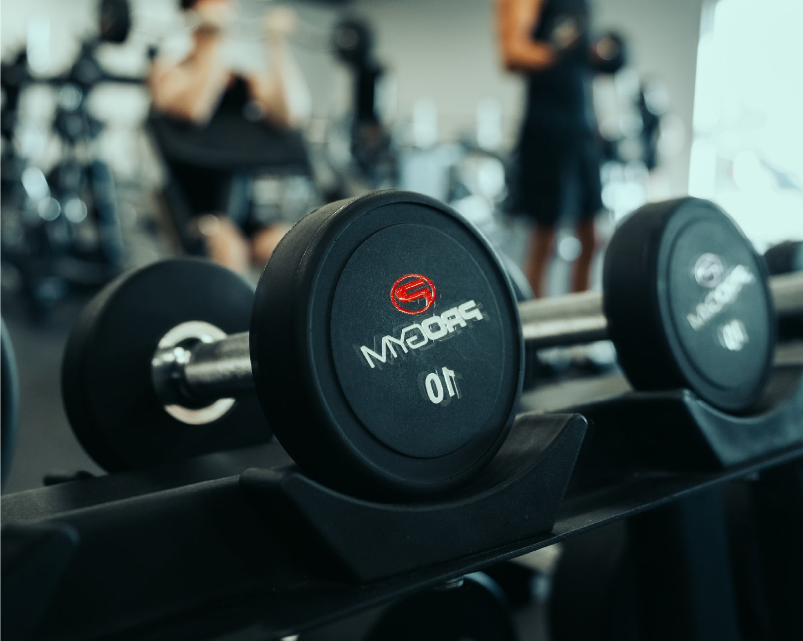 Haltère noire de 10 lb avec logo ProGym, posée sur un support dans une salle d’entraînement, avec des personnes s’entraînant en arrière-plan flou.