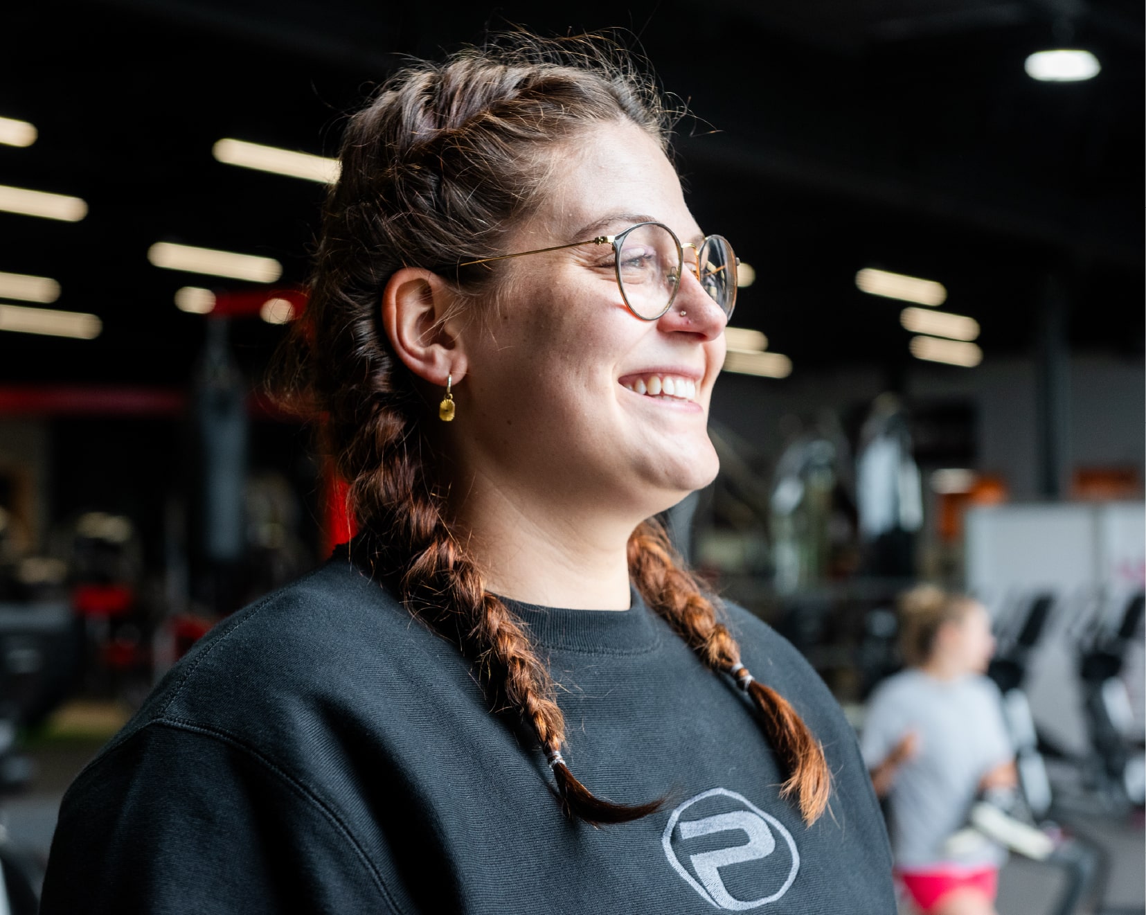 Femme souriante portant des lunettes rondes et un chandail noir ProGym, les cheveux tressés, dans une salle d’entraînement à l’ambiance conviviale.