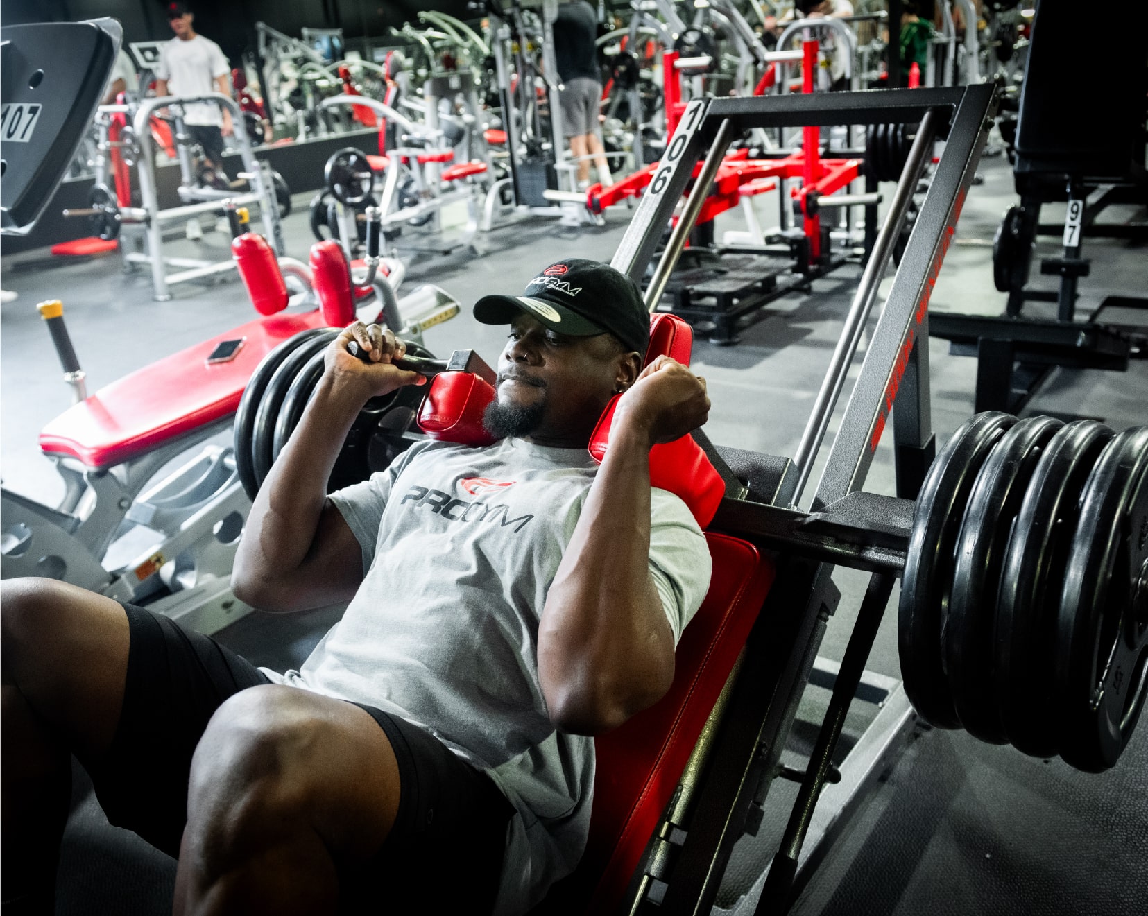 Homme concentré effectuant un exercice de presse à jambes sur une machine rouge et noire, vêtu d’un t-shirt gris et d’une casquette noire ProGym, dans une salle de musculation bien équipée.