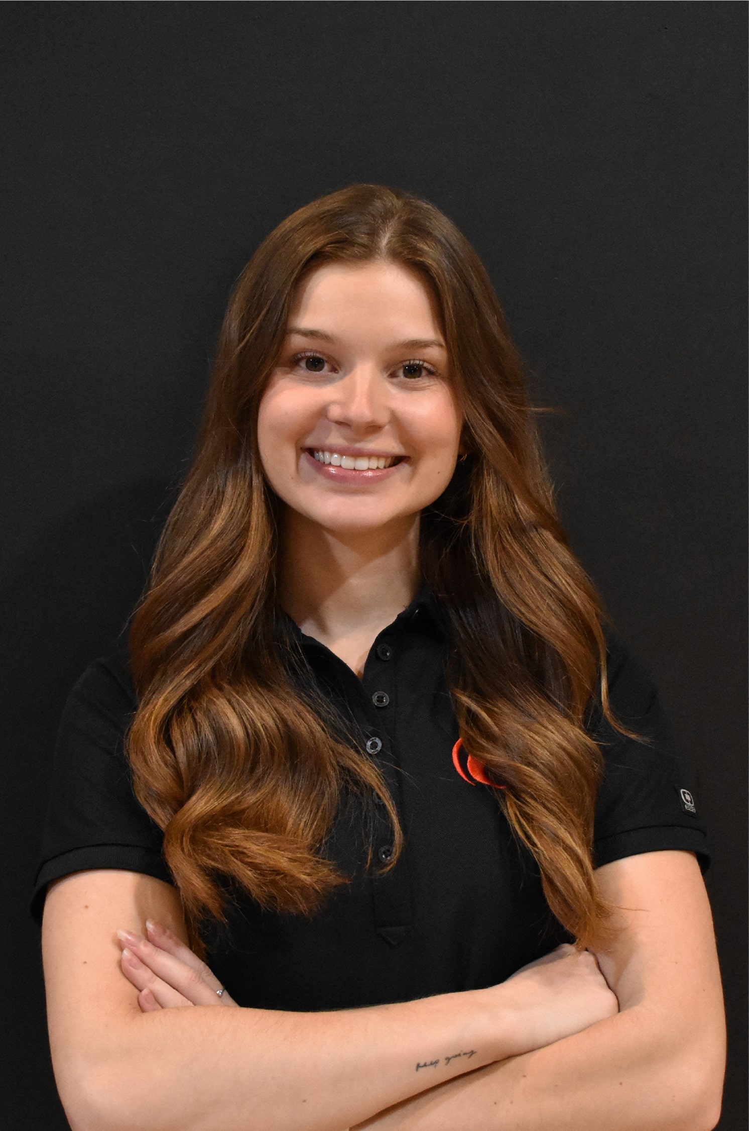Emie, souriante, portant un polo noir au logo rouge de ProGym, bras croisés devant un mur noir.