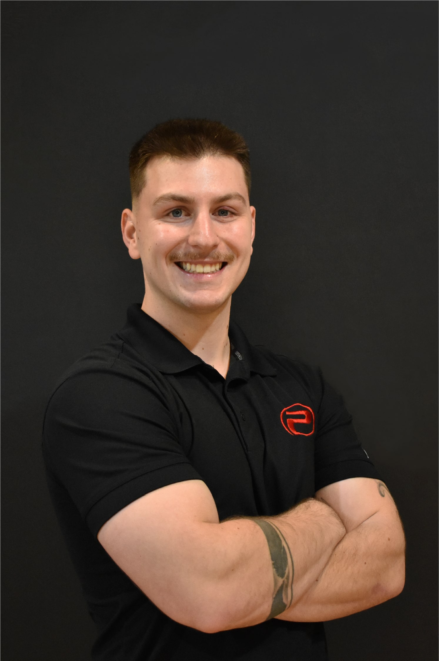Gregory, souriant, portant un polo noir au logo rouge de ProGym, bras croisés devant un mur noir.