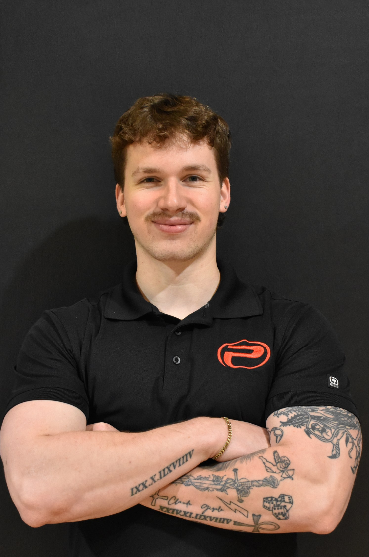 Isaak, bras croisés, portant un polo noir avec le logo rouge de ProGym, debout devant un mur noir.