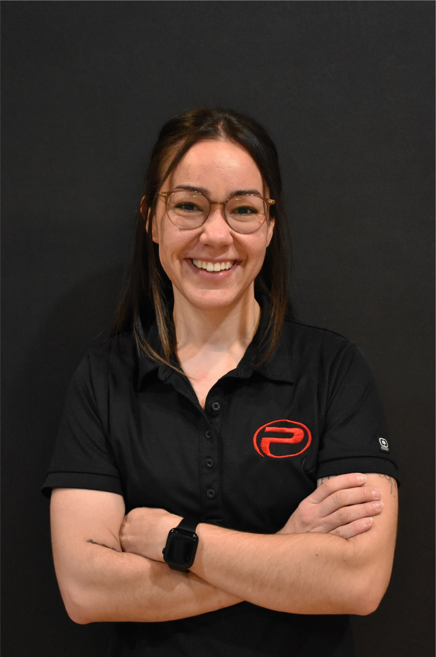 Noémie, souriante, bras croisés, portant un polo noir avec le logo rouge de ProGym, debout devant un mur noir.