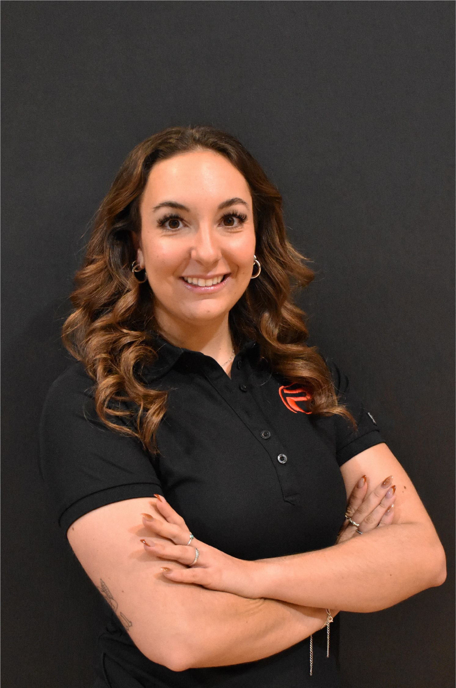 Noémie, souriante, bras croisés, portant un polo noir avec le logo rouge de ProGym, debout devant un mur noir.