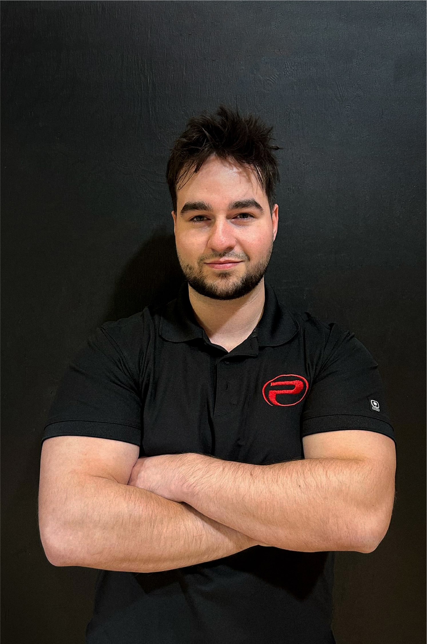 Samue, bras croisés portant un polo noir avec le logo rouge de ProGym, debout devant un mur noir.